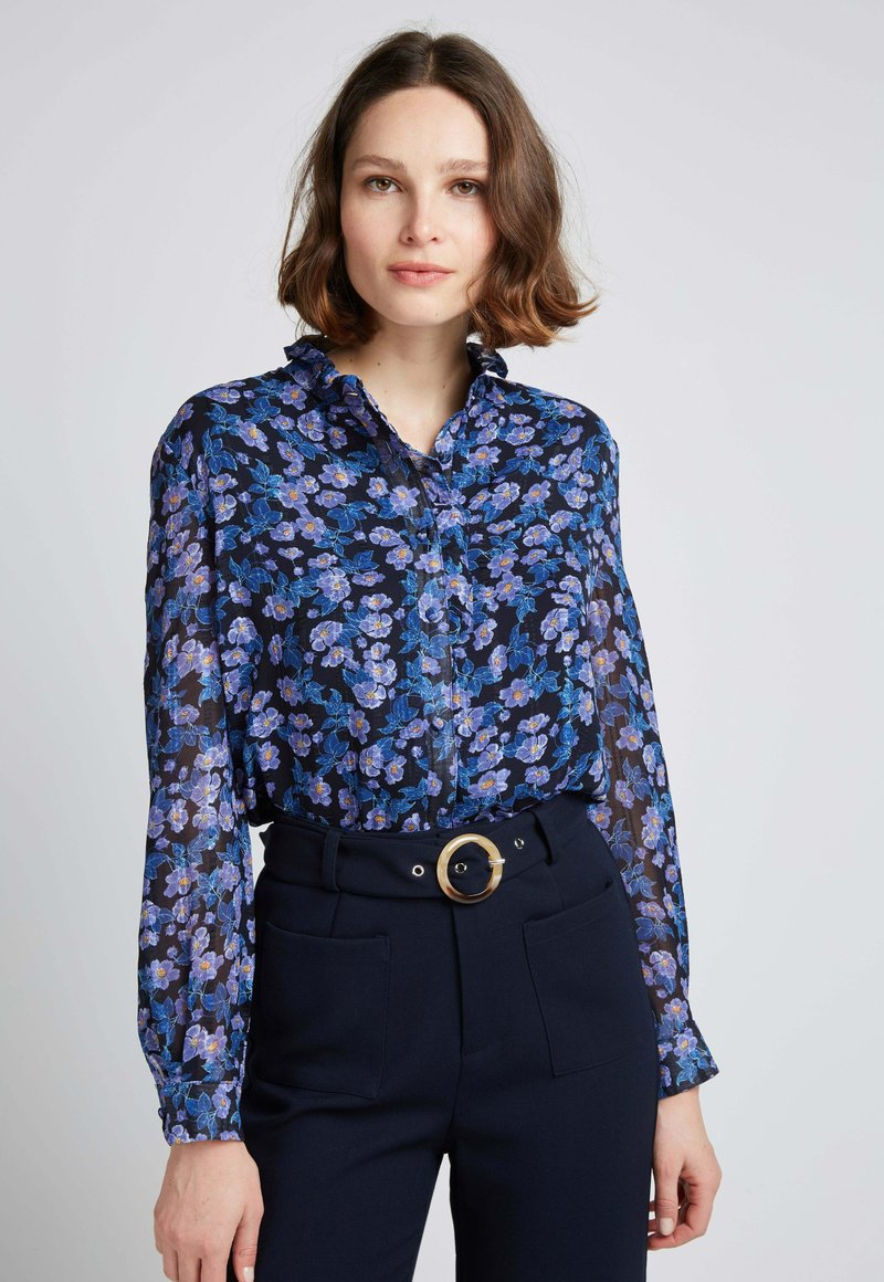 Chemisier à manches longues et boutonné, de couleur bleu marine, avec un motif floral dans des tons de bleu et de violet, doté d'un col et de poignets à volants.