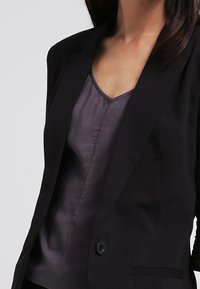 Sort skræddersyet blazer med en glat tekstur, enkelt knaplukning og klapkraver, lagd over en mørk satin camiskjorte.