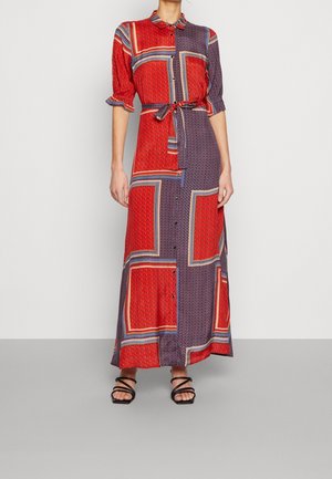 Kobieta nosząca długą sukienkę na guziki z czerwonymi i fioletowymi geometrycznymi wzorami blokowymi oraz czarne sandały na paski.