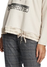 Beige långärmad topp med glittrande text "SKAPATLYCKAN" och satingall, i kombination med blå rutiga byxor och synlig hand.