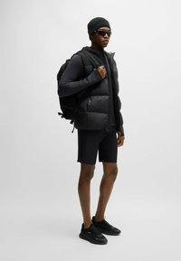 Doudoune noire portée sur un tee-shirt à manches longues noir, associée à un short noir et des baskets noires. Les accessoires comprennent un bonnet noir et des lunettes de soleil.