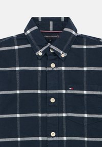 Camisa quadriculada azul-marinho com padrão de grelha branca, colarinho abotoado, seis botões, pequeno bolso à esquerda do peito e logótipo bordado da bandeira.