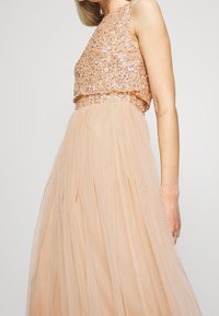 Pitkä persikkamekko, jossa on paljettikoristeltu pusero, pyöreä pääntie ja virtaava hame, joka on valmistettu tyllistä. Puseroa on koristeltu lisäyksillä, jotka tuovat lisää yksityiskohtia ja tekstuuria.