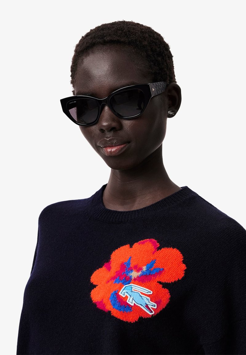 Schwarze Cateye-Sonnenbrille mit getönten Gläsern, getragen mit einem navyblauen Pullover, der mit einem großen rot-orangefarbenen floralen Stickdesign verziert ist.