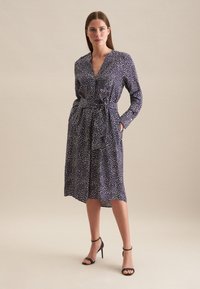 Robe bleu marine à pois blancs, manches longues et ceinture à la taille. Elle présente un décolleté en V et des poches latérales. Porter avec des talons noirs.