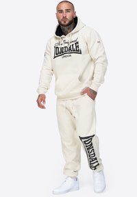 Krämfärgad hoodie med svart textlogotyp, tillsammans med matchande joggers med logotryck. Vita sneakers fulländar outfiten.