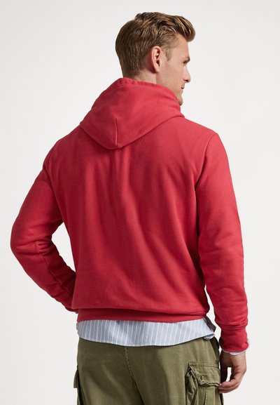 Muškarac s kratkom plavom kosom obučen u crvenu hoodicu, plavu prugastu majicu i zelene cargo hlače, okrenut leđima, na jednostavnoj pozadini.