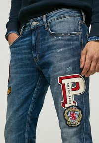 Jean en denim bleu foncé avec des zones abîmées, ornés de grandes broderies, y compris une lettre "P" rouge et un logo circulaire.