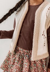 Kremowa kamizelka z polaru z brązowymi wykończeniami noszona z brązową bluzką z długim rękawem, w połączeniu z zwiewną spódnicą w kwiaty w kolorach czerwonym, białym, turkusowym i brązowym.