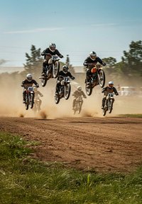 Six motos, avec des pilotes en combinaisons noires et casques, sautent à travers la terre, créant des nuages de poussière. Les motos affichent des couleurs variées et des numéros de course.