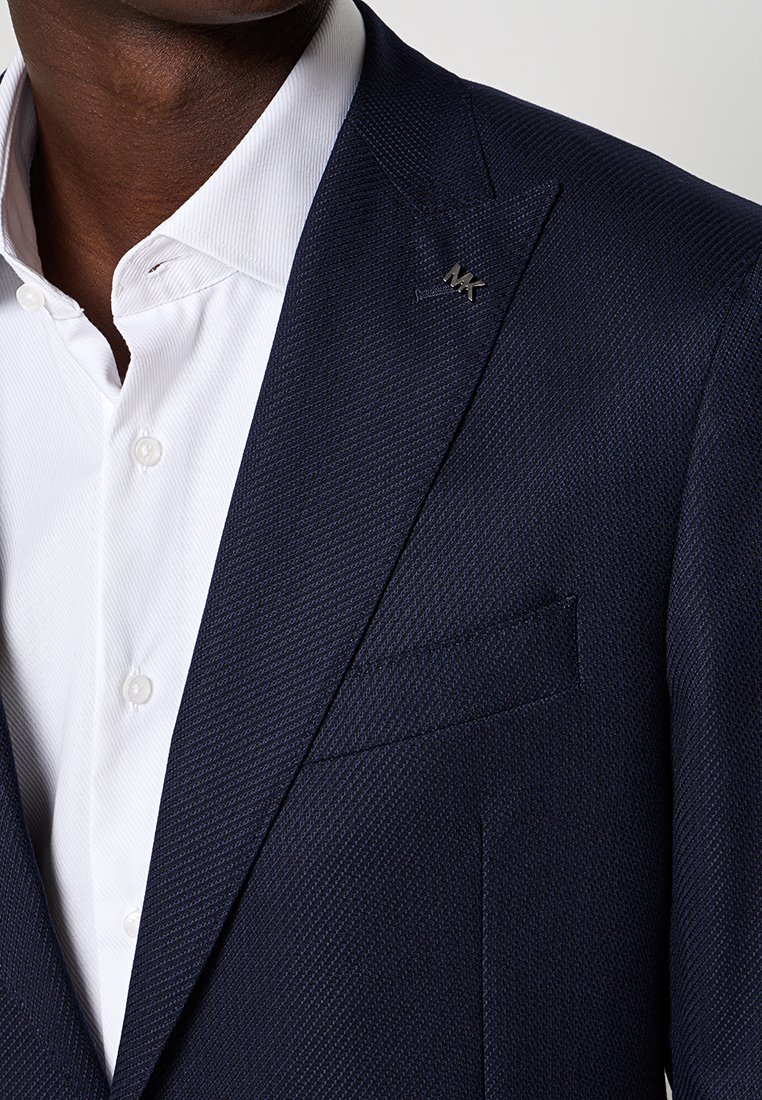 Marineblaue Blazer aus strukturiertem Stoff, mit einem Reverskragen, Tasche und MK-Logo-Anstecker am Kragen, getragen über einem weißen Hemd.