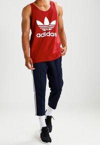 Červený bavlněný tílko s bílým logem Adidas a grafikou, spárované s námořnickými tepláky s bílými bočními pruhy a černými teniskami.