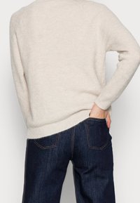 Pull en tricot beige avec texture côtelée, encolure ronde et fente sur le côté. Associé à un jean bleu foncé avec des surpiqûres contrastées.