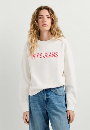 Jeune femme aux longs cheveux ondulés portant un sweat-shirt blanc avec le texte rouge « PEPE JEANS » et un jean bleu, debout devant un fond clair uni.