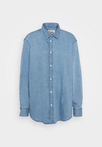 Camisetta in denim azzurro chiaro a maniche lunghe, con colletto a punta e bottoni snap bianchi, esposta su uno sfondo bianco.