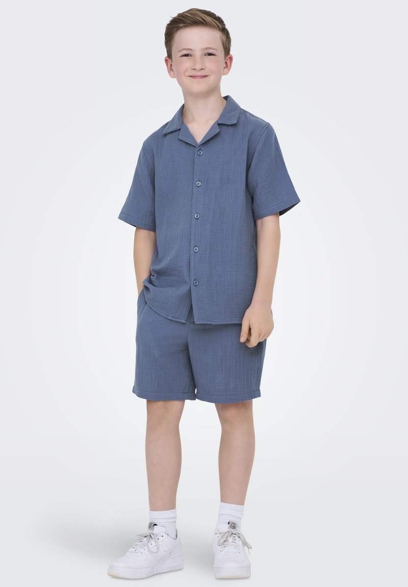 Blå kortärmad skjorta och matchande shorts i strukturerad tyg. Har en krage, knappstängning och sidofickor. Bärs med vita sneakers.
