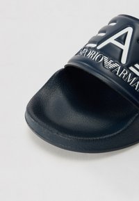 Morski drsni sandal s teksturirano gumirano zgornjimi deli, z belim logotipom in konkavnimi črkami na traku, odprtimi prsti in oblikovanim podplatom.