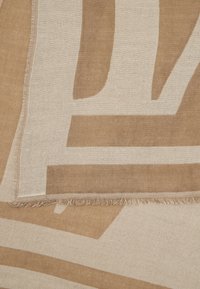 Beige og kremfarget vevd tekstil med geometriske mønstre og frynser i kantene, brettet for å vise lagdelt tekstur og design.