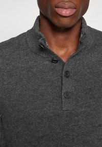 Nahaufnahme einer Person, die einen dunkelgrauen Strickpullover mit einem Knopfkragen trägt, der mit vier schwarzen Knöpfen ausgestattet ist, von denen zwei geschlossen und zwei offen sind.