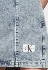 Denimkjol i ljusblått med en sliten textur, som har en sydd söm och en vit etikett med "Calvin Klein Jeans"-logotypen vid nederkanten.