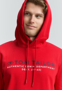 Jeune homme portant un sweat à capuche rouge avec le texte "TOM TAILOR Authentic Denim Department", regardant sur le côté avec un léger sourire.