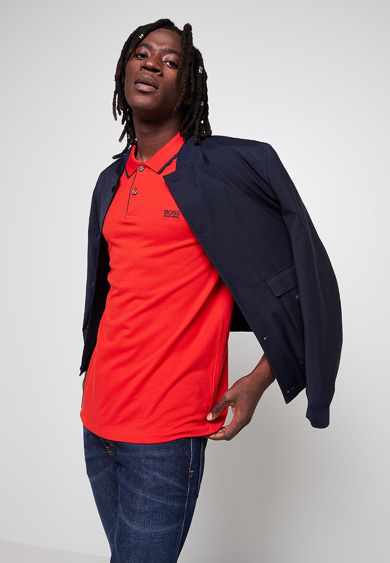 Polo rouge avec trois boutons et logo brodé, superposé sous une veste marine, porté avec un jean en denim bleu. Individu sur un fond neutre.