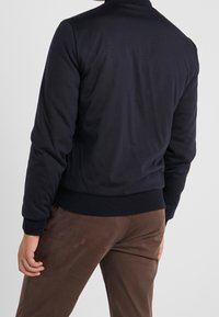 Giacca bomber blu navy con colletto e polsini a costine, caratterizzata da una texture liscia e un design aderente, abbinata a pantaloni marroni.