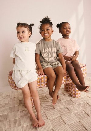 Három kisgyermek ül egy kockás padon, laza sortot és grafikus mintás pólót viselnek, mosolyognak és különböző irányokba néznek.