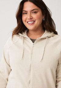 Beige Zip-Hoodie mit strukturierter Oberfläche, Kordelzugkapuze und silbernem Reißverschluss. Verfügt über Raglanärmel für eine lockere Passform.