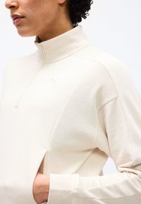 Άτομο που φορά ανοιχτόχρωμο μπεζ πουλόβερ με φερμουάρ έως τη μέση, με υφασμάτινες λεπτομέρειες και μικρό λογότυπο στο στήθος, με το χέρι τσουγκρισμένο στην πλαϊνή τσέπη.