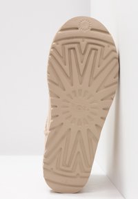 Semelle de bottes en caoutchouc beige avec motif de bande de roulement en zigzag profond et motif en forme de rayons de soleil, affichant le nom de la marque « UGG » au centre.