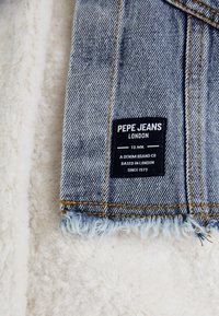 Lyseblåt denimstof med frynset kant, der har et sort mærke med teksten "PEPE JEANS LONDON." Synlig gul syning langs sømmene.
