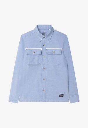 Chemise bleu clair à manches longues avec un col boutonné, deux poches poitrine, des accents blancs, et un patch logo Timberland noir à l'ourlet.