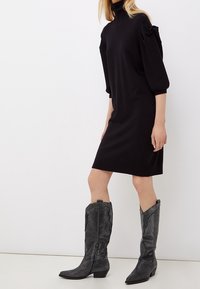 Vestito nero alla lunghezza del ginocchio con colletto alto e maniche a sbuffo; abbinato a stivali texani neri con texture, caratterizzati da tacco basso e punta affilata.