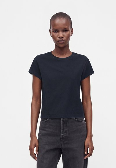 Jeune personne debout face à l'avant portant un t-shirt noir uni à manches courtes et un jean gris foncé sur fond blanc.