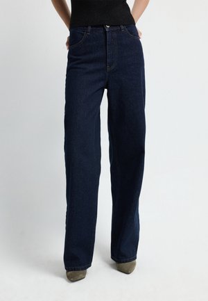 Jean bleu foncé taille haute à jambes larges porté avec un haut noir et des chaussures beiges pointues, mains du mannequin dans les poches devant un arrière-plan uni.