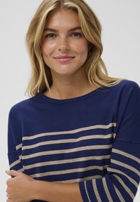 Marineblauer Pullover mit beigen horizontalen Streifen, Rundhalsausschnitt, gerippten Bündchen und weichem, strukturiertem Stoff. Modell mit verschränkten Armen, lächelnd.