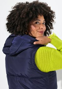 Marinblå vadderad väst med huva, med en arm i klargrön knuten stickning. Tyget verkar slätt och glänsande, vilket framhäver kontrasterande texturer.