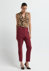 Woman wearing sleeveless leopard print top, high-waisted maroon tailored pants, and black high-heeled shoes, standing against white background.