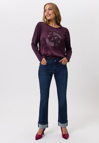 Langsleeve bordeaux top met een cirkelvormig grafisch ontwerp in strass, gecombineerd met donkerblauwe jeans die aan de manchetten zijn opgerold, en roze hakken.
