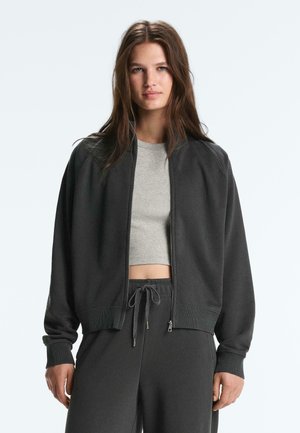 Tânără cu păr lung și castaniu, purtând o jachetă neagră cu fermoar, top scurt gri și pantaloni negri cu șnur, pe un fundal simplu.