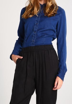 Vrouw draagt een blauwe denim blouse met knopen, ingestopt in hoge, losvallende zwarte broek, met één hand in de zak.