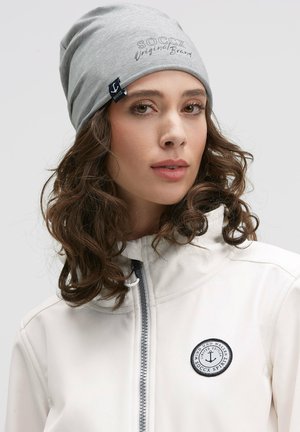 Gray beanie with embroidered "SOCCX Original Brand" and navy tag, paired with a white zip-up jacket featuring a round black patch.