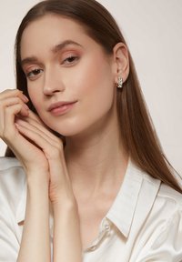 Goldene Creolen mit strukturierter Oberfläche, die klare Akzente zeigen. Modell mit langen, glatten Haaren, das ein weißes Satinhemd trägt. Die Hände sind elegant positioniert.