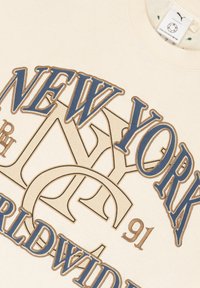 Crème-kleurige sweatshirt met grote teksten "NEW YORK" en "WORLDWIDE" in marineblauw en tan. Zachte stof met een ronde hals en zichtbare label.