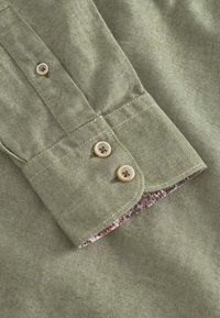 Camicia verde oliva con texture a righe. Presenta un polsino con bottone e un dettaglio floreale all'interno del colletto. Due bottoni visibili.