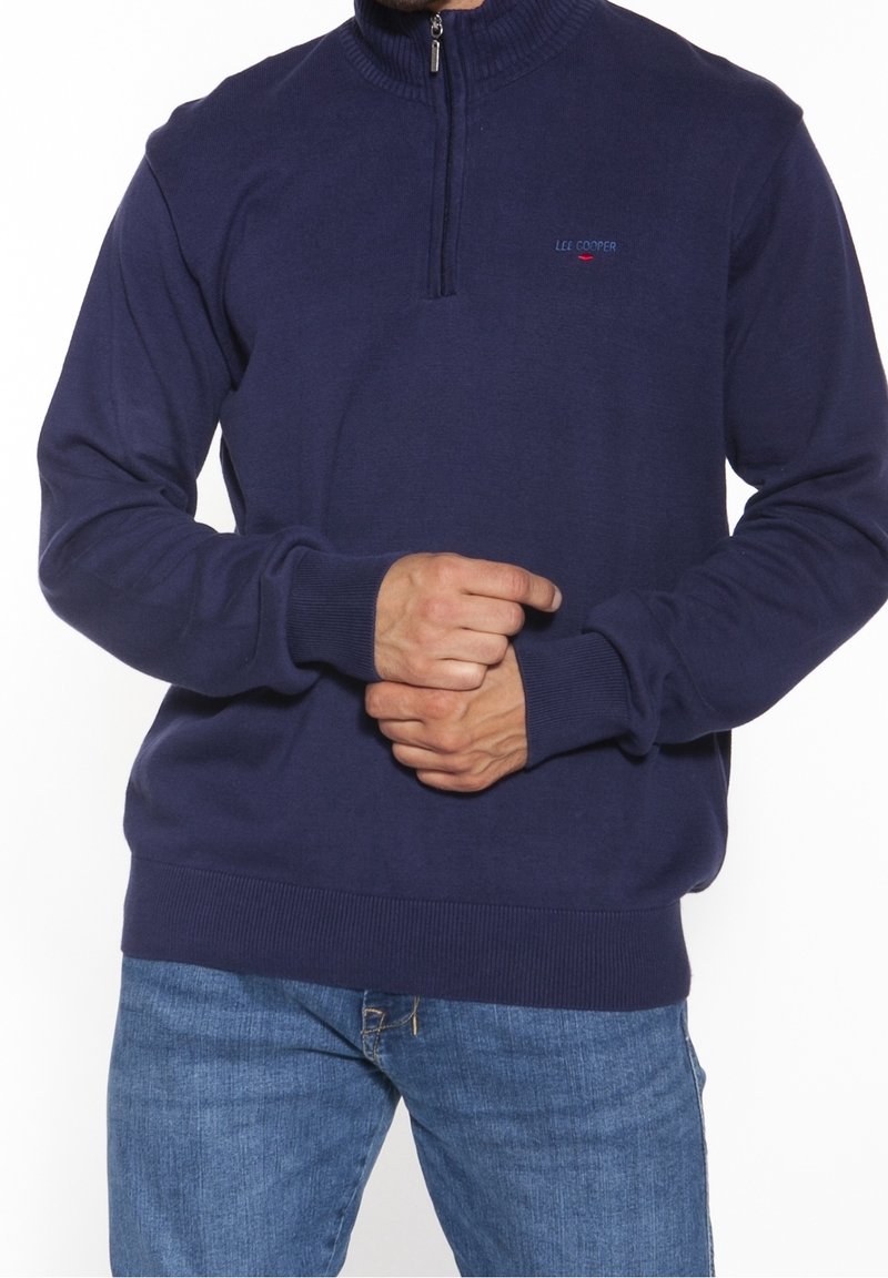 Mężczyzna ubrany w granatowy sweter na zamek do połowy kołnierza z prążkowanymi mankietami i krawędzią, w połączeniu z niebieskimi dżinsami, z rękami splecionymi przed sobą.