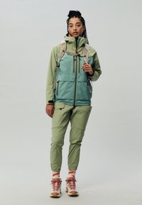Giacca verde leggera con cappuccio e molte tasche, abbinata a pantaloni tapered coordinati. Sneakers marroni e rosa completano l'outfit.