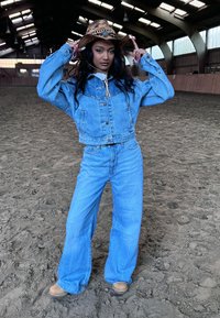 Veste en denim et jeans à jambes larges en délavé clair, avec un chapeau en paille marron orné d'accents turquoise, dans une arène intérieure avec un sol en sable.