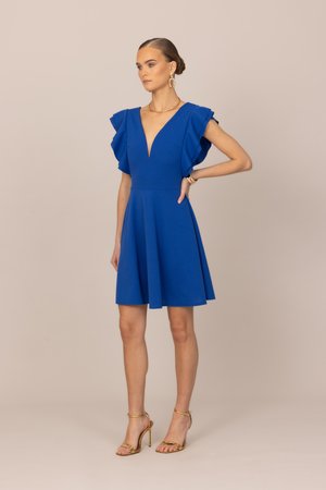 Kvinde iført en blå knælængde kjole med flæseærmer og V-hals, gyldne hoop-øreringe, halskæde, armbånd og gyldne højhælede sko.